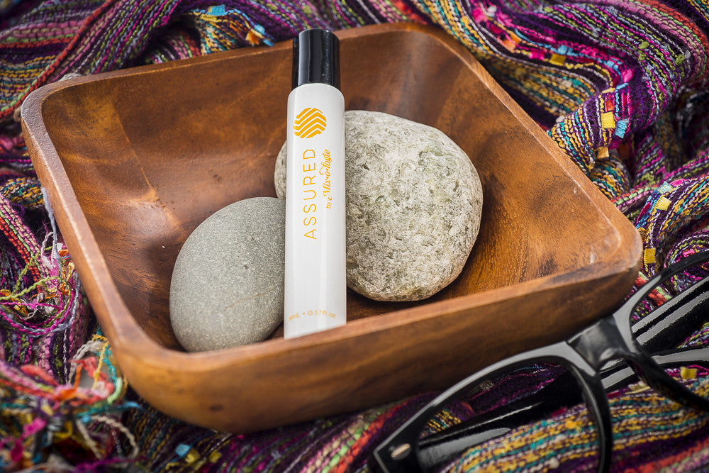 Assured (Natural) white cylinder rollerball with mustard color lettering. Rollerball has 0.17 fl oz or 5 mL. Rollerball pictured in wood container with rocks on a colorful blanket.