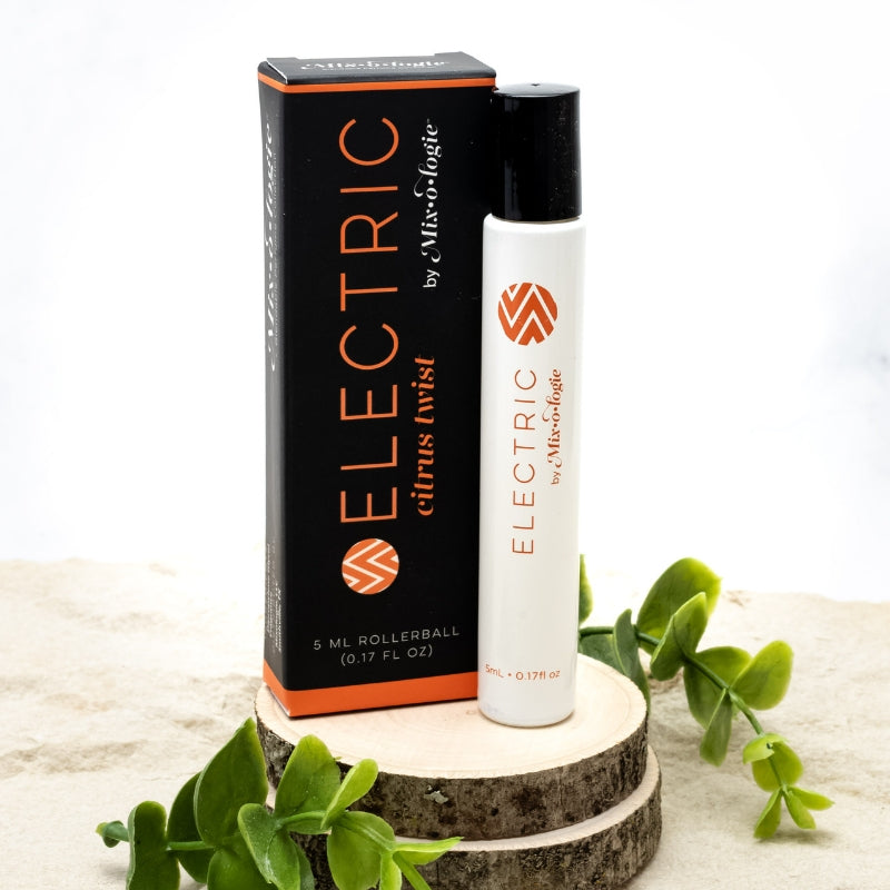 Electric (Citrus Twist) white cylinder rollerball with orange color lettering with black box and orange color lettering. Rollerball has 0.17 fl oz or 5 mL. Rollerball and rollerball box pictured on wood in the sand with greenery.